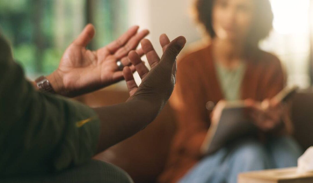 A close-up, shallow depth-of-field shot focusing on a client's expressive hand gestures with open palms during a discussion. In the blurred background, a female therapist sits on a couch holding a notebook and pen, listening attentively in a warm, softly lit office environment.