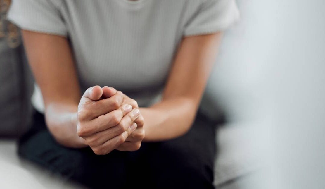 A close-up, shallow depth-of-field shot focusing on a person's hands tightly clasped together, conveying anxiety, stress, or emotional tension. The person is wearing a white t-shirt and black pants while sitting on a grey couch, with their face cropped out and the background softly blurred.