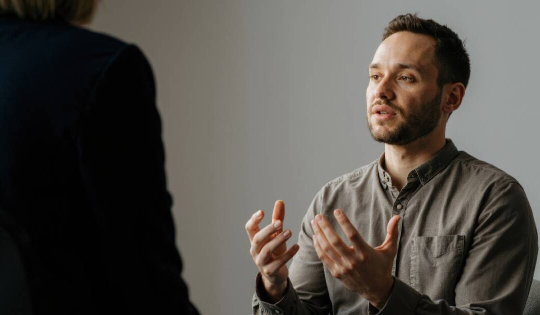 An over-the-shoulder shot of a man with short dark hair and a beard, wearing a grey button-down shirt. He looks ahead with an earnest expression, gesturing with both hands as he speaks. The dark silhouette of an interviewer or counselor is visible in the foreground against a plain, neutral background wall.