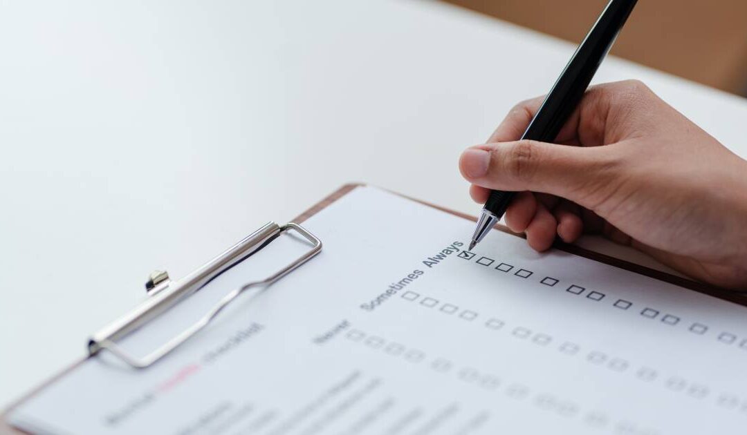 A close-up shot of a person's hand holding a black pen to check off a box on a survey or assessment form. The document is secured to a wooden clipboard on a white table, featuring response columns labeled "Never," "Sometimes," and "Always," with a shallow depth of field blurring the lower part of the page.