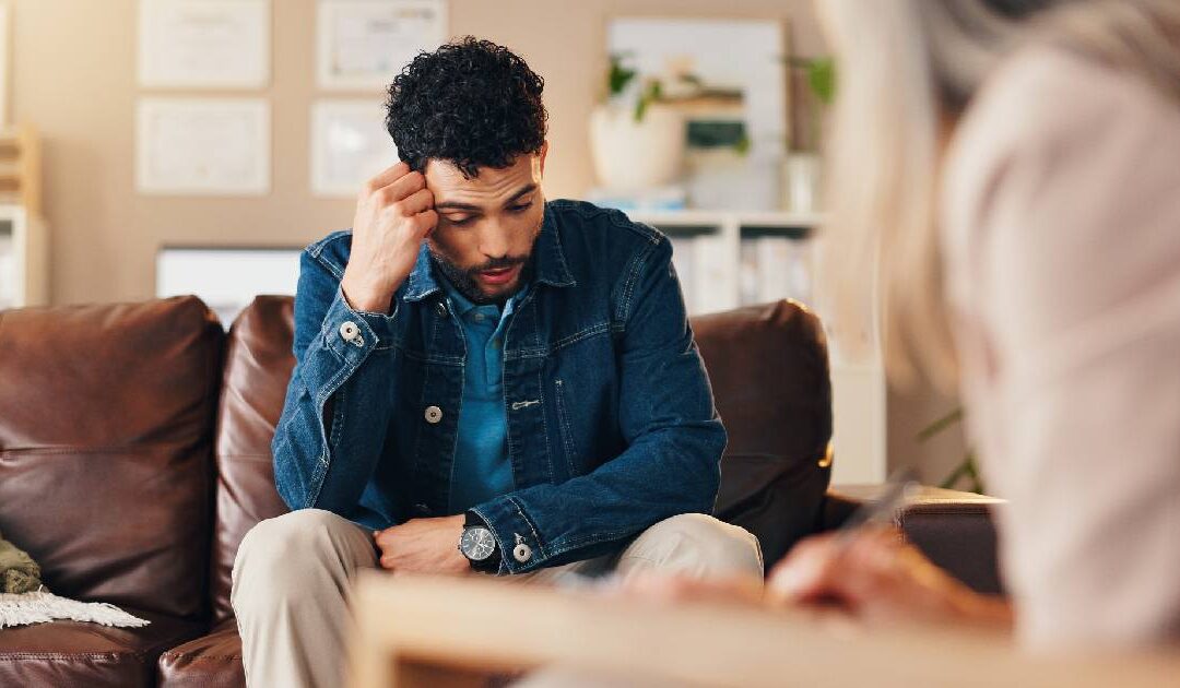 An over-the-shoulder shot of a distressed young man with curly dark hair and a beard during a counseling session. He is sitting on a brown leather couch wearing a denim jacket and khaki pants, holding a hand to his temple with a worried, downward expression. In the blurred foreground, a therapist is actively taking notes.