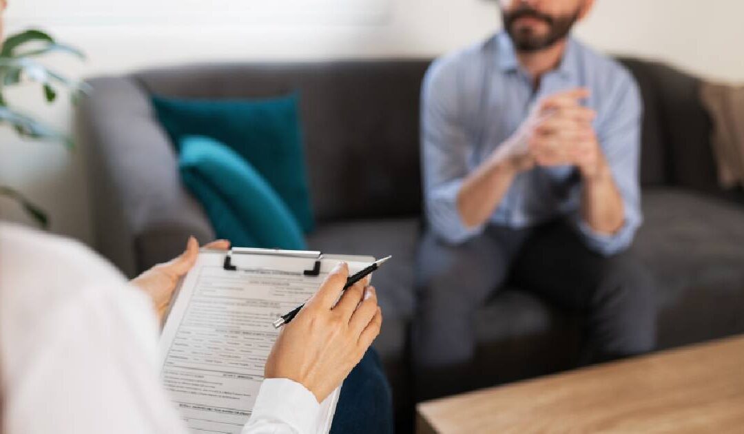 An over-the-shoulder shot focusing on a female therapist's hands holding a black pen and a clipboard with an intake or assessment form. In the blurred background, a bearded man in a light blue button-down shirt sits on a dark grey sofa with clasped hands, participating in a professional consultation.