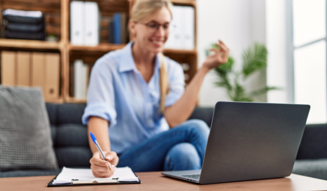 A close-up shot focusing on an open laptop and a hand writing with a blue pen on a clipboard. In the softly blurred background, a smiling blonde woman wearing glasses and a light blue shirt sits on a grey sofa, gesturing during a virtual meeting or video call in a bright office space.