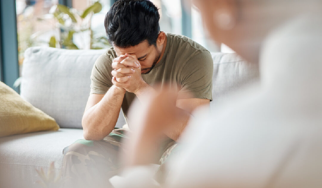 An over-the-shoulder shot focusing on a distressed man wearing an olive green t-shirt and camouflage pants during a therapy session. He sits on a light grey couch with his eyes closed and hands tightly clasped to his forehead in deep emotional distress. In the blurred foreground, a counselor listens attentively in a bright, sunlit room.